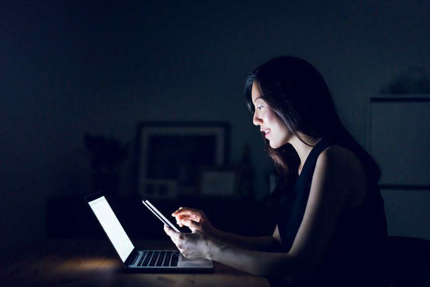 Side view of Asian woman working remote from both a smart device and laptop